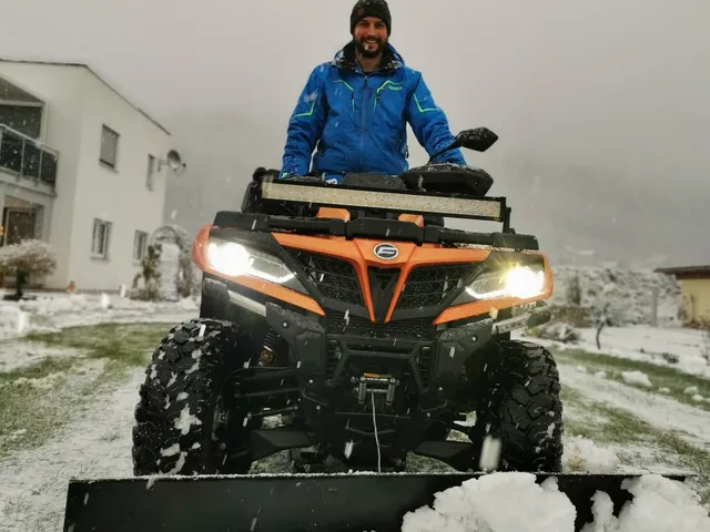 Ein Mann sitzt auf einem orangefarbenen ATV mit Schneepflug und räumt Schnee. Es schneit und im Hintergrund ist ein Haus zu sehen. 
