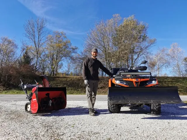 Ein Mann steht neben einer Honda-Schneefräse und einem orangefarbenen ATV mit Schneepflug. Der Himmel ist blau und es gibt Bäume im Hintergrund
