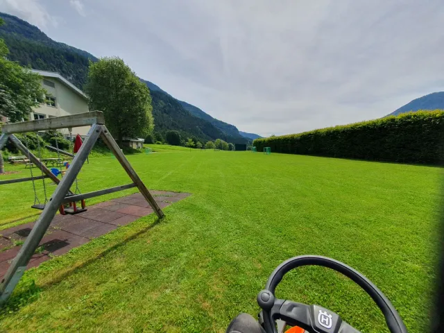 "Ein Rasenmäher fährt über eine grüne Wiese mit einem Spielplatz und Bergen im Hintergrund. Die Landschaft ist sonnig und die Wiese ist gepflegt.